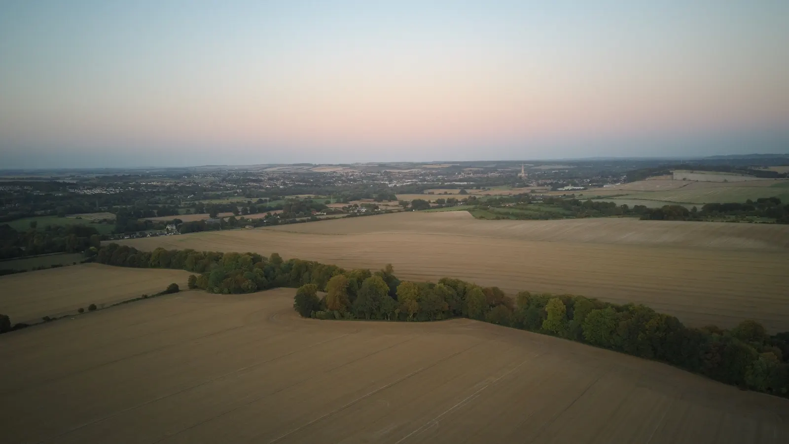 Aerial view at dusk over golden harvest fields near Salisbury, with the city and cathedral spire on the horizon under a pink sky