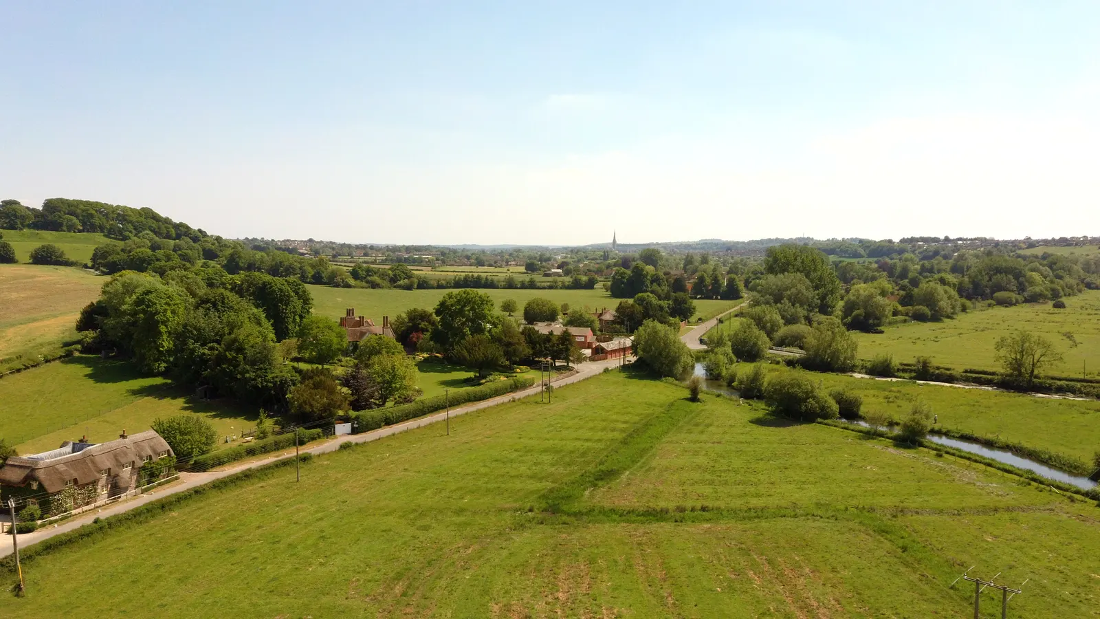 Aerial summer view of a Wiltshire river valley near Salisbury — water meadows, a winding lane, a thatched cottage and the cathedral spire in the far distance